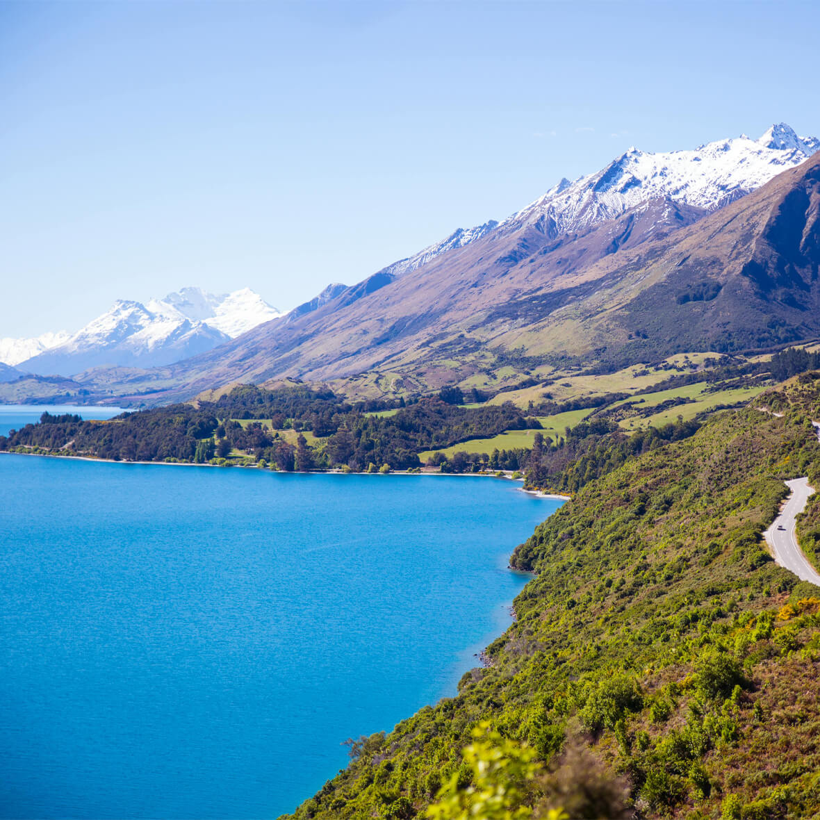 A scenic view of a vibrant turquoise lake bordered by verdant hills. Snow-capped mountains rise in the background under a clear blue sky. A winding road follows the contours of the hills along the lake's edge.