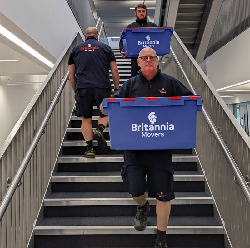 Three movers from Britannia Movers are carrying large blue containers labeled with the company's logo up a staircase. The two movers in the front are ascending, while the mover in the back is also on the stairs. All are wearing black uniforms.