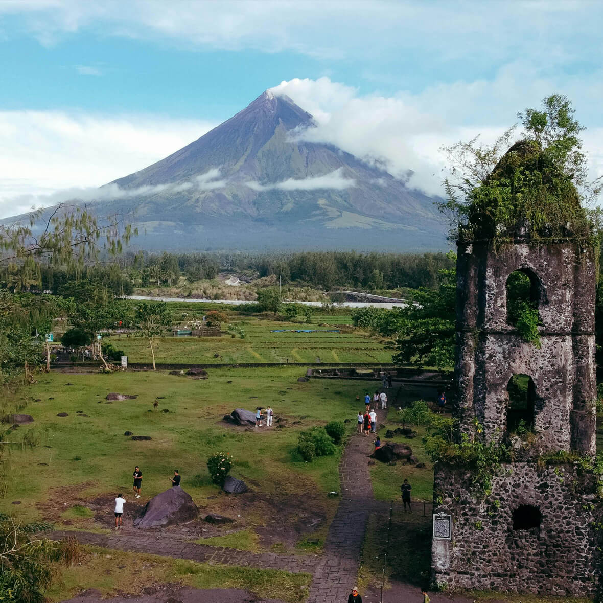 A scenic view of Mayon Volcano in the Philippines, featuring its symmetrical cone shape and a partly cloud-covered peak. In the foreground is Cagsawa Ruins, with people walking around the grassy area and the stone belfry tower standing prominently.