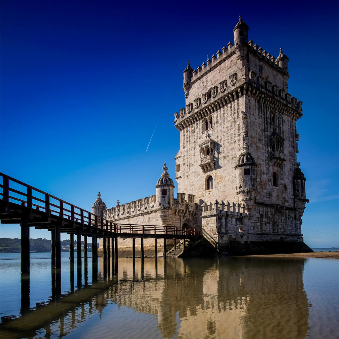 A stone tower with ornate turrets stands on the waterfront, connected to land by a wooden bridge. The structure is reflected in the calm water below, and the sky overhead is clear and blue with a contrail cutting across it.