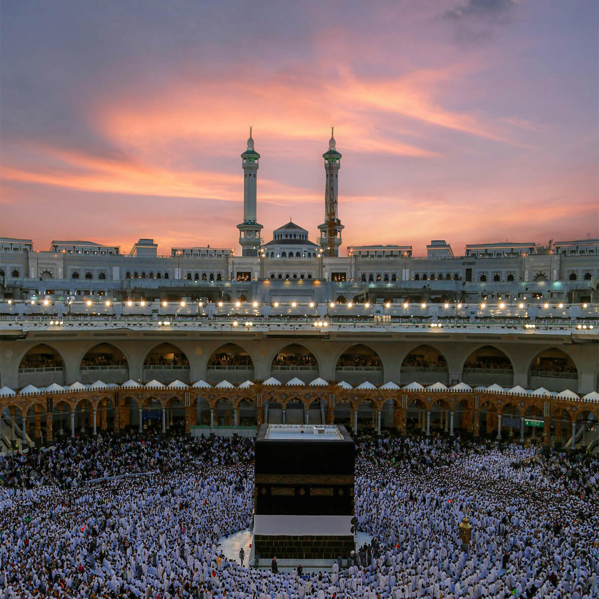 Thousands of pilgrims gather around the Kaaba at Masjid al-Haram in Mecca, Saudi Arabia during dusk. The sky is painted with shades of pink and orange as the evening sets in. Two minarets stand tall in the background, and the mosque is illuminated with lights.