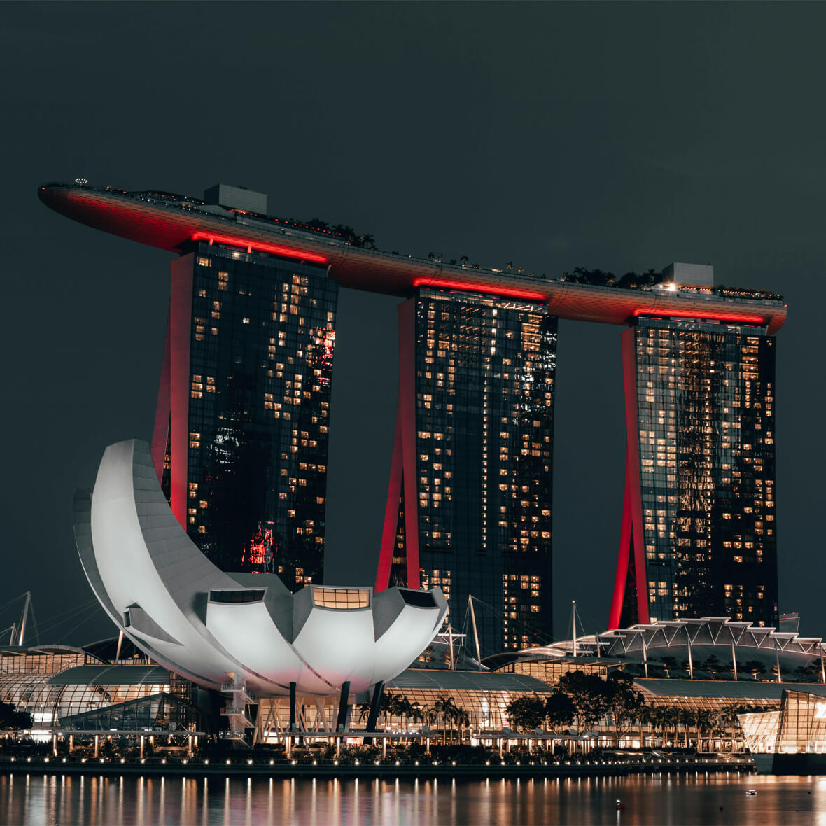 Night view of Singapore's Marina Bay Sands, a prominent hotel featuring three towers connected by a rooftop SkyPark with an infinity pool. In the foreground, a lotus-like structure, the ArtScience Museum, stands illuminated against the night sky.