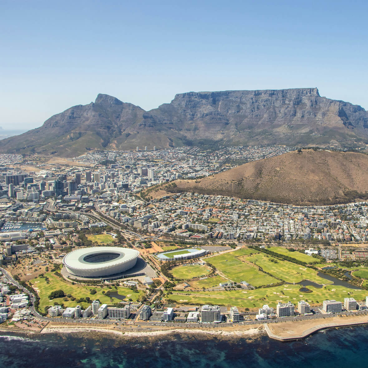 Aerial view of Cape Town, South Africa, showcasing the Cape Town Stadium near the shoreline. The cityscape is nestled between the coastal waters and the iconic flat-topped Table Mountain in the background. Lion's Head mountain is also visible to the right.