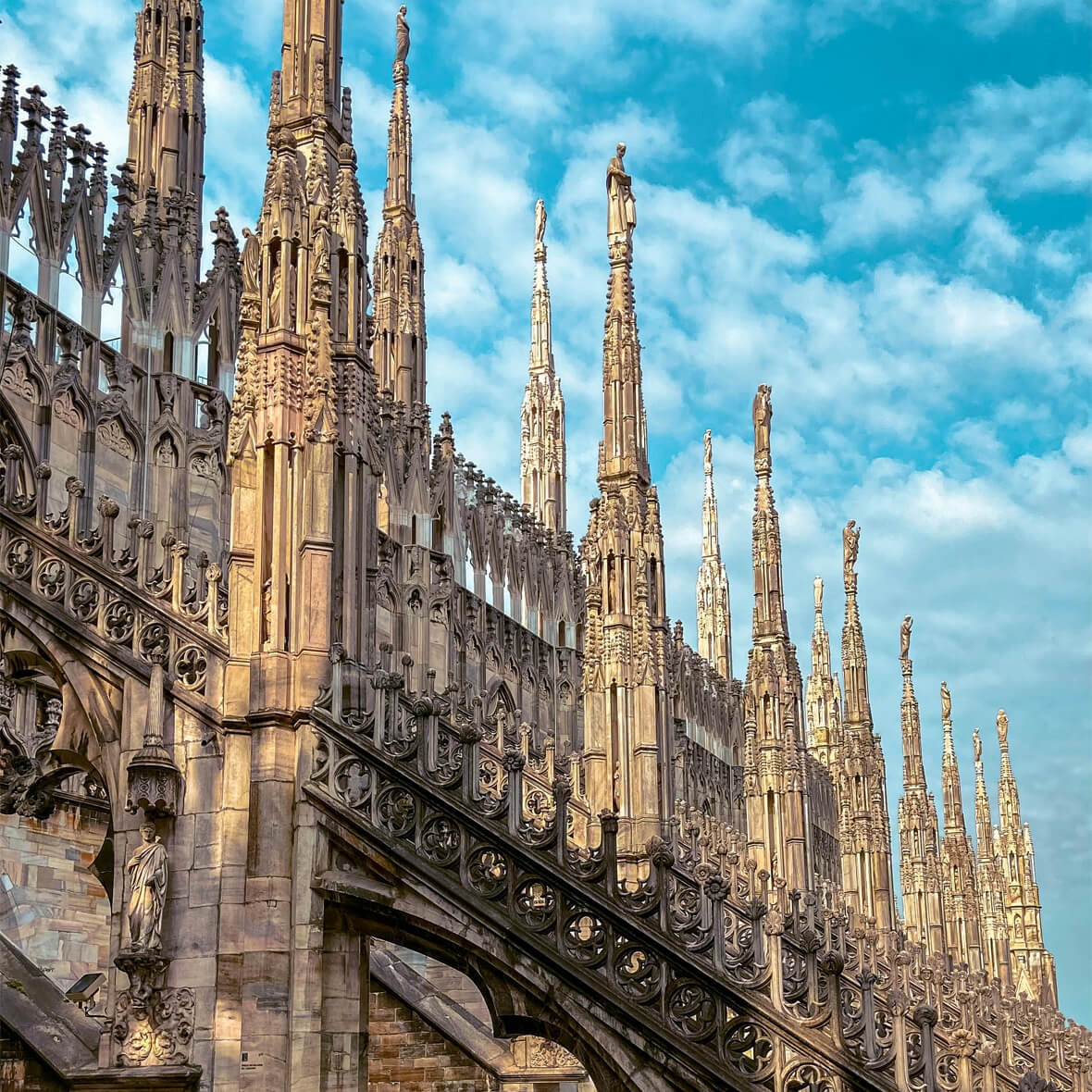 Exterior view of the intricate spires of Milan Cathedral (Duomo di Milano) against a blue sky with scattered clouds. The Gothic architecture is characterized by detailed carvings, pointed arches, and flying buttresses.