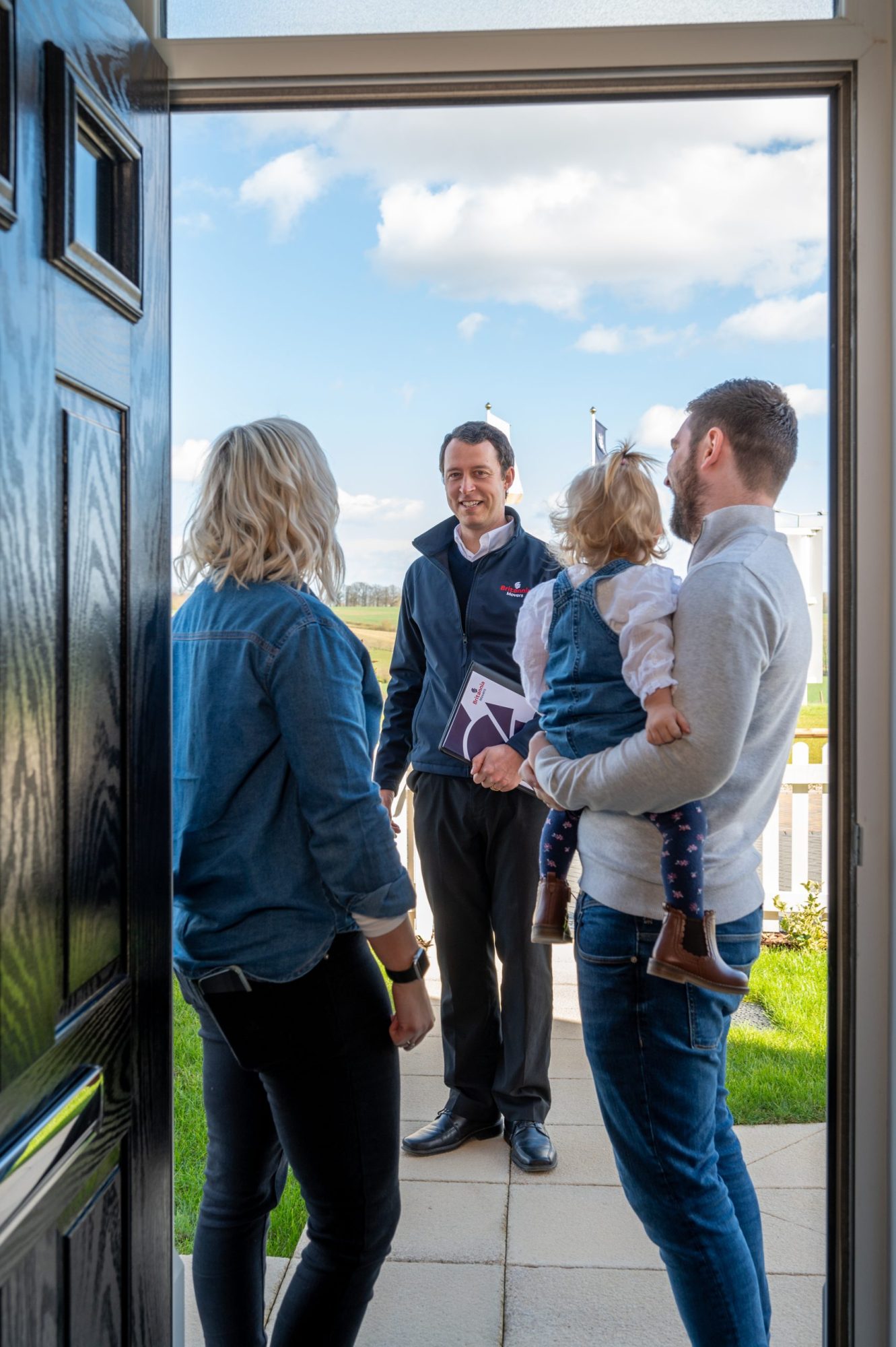 A man holding a child and a woman stand at the threshold of their front door, conversing with a man in a jacket who is holding brochures. The scene appears to be a pleasant day with blue skies and white clouds in the background.