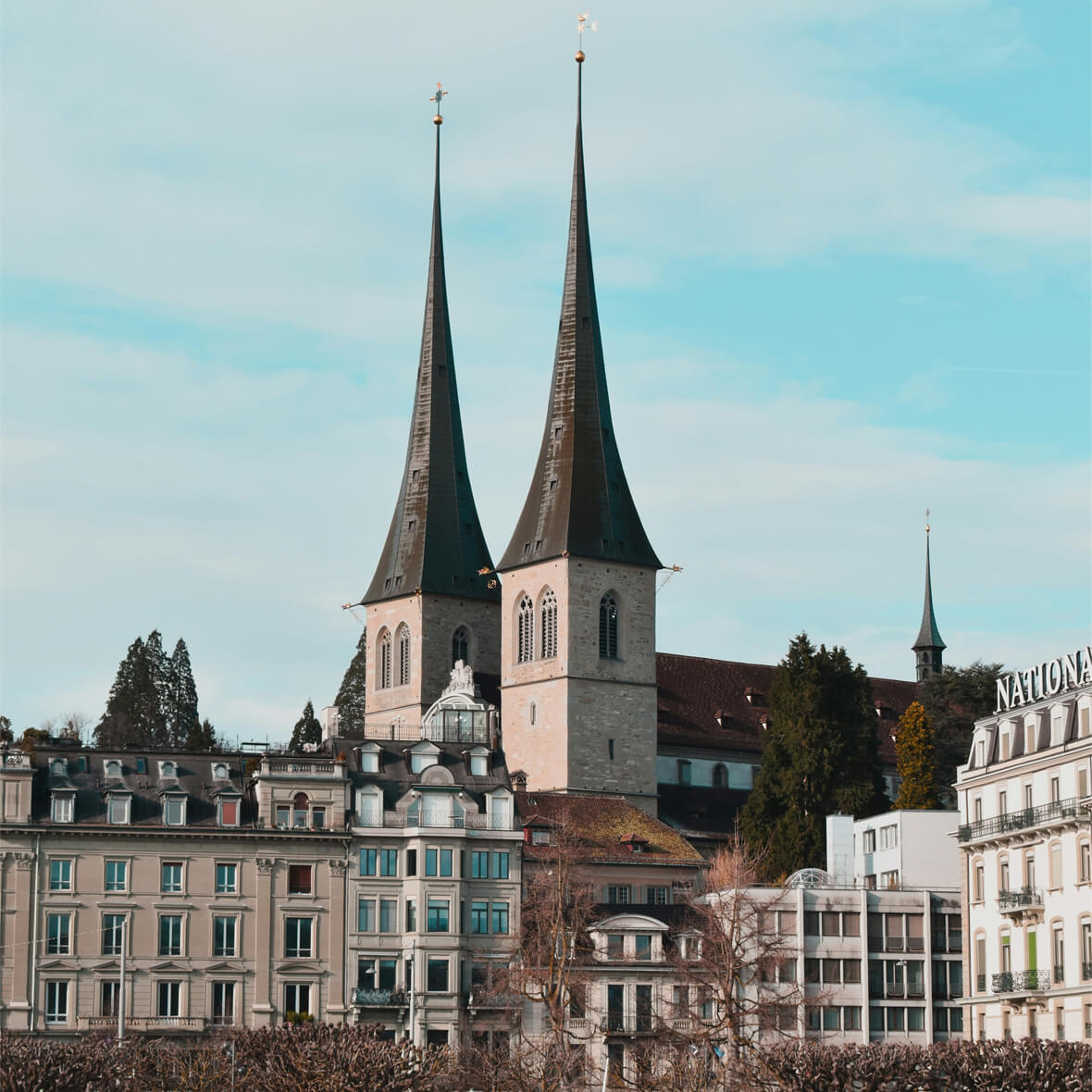 A scenic view of a European city featuring a historic church with two tall spires rising above residential and commercial buildings. The sky is clear with a few clouds, and the architecture showcases a blend of traditional and modern styles.