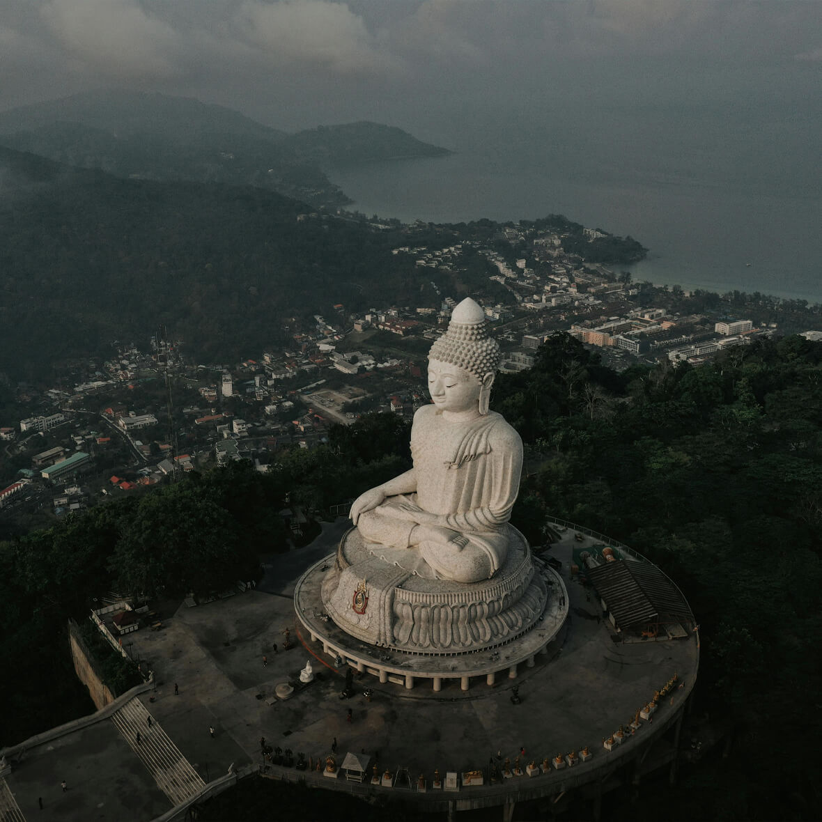 Aerial view of a large white Buddha statue seated on a circular platform overlooking a town and coastline surrounded by lush green hills under a cloudy sky. The town's buildings and coastal line are visible in the background.