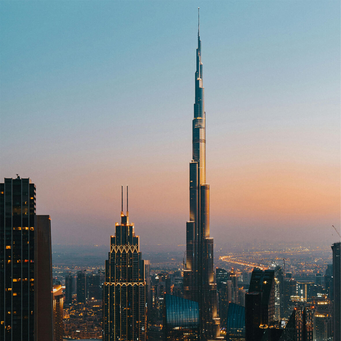 A tall skyscraper, the Burj Khalifa, stands prominently against a dawn or dusk sky in a cityscape filled with other illuminated buildings. The scene captures the towering structure amidst the surrounding urban environment.