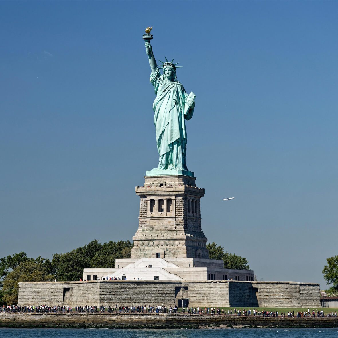 The Statue of Liberty stands tall on Liberty Island against a clear blue sky. Crowds of visitors are seen at the statue's base. The iconic statue holds a torch in her right hand and a tablet in her left, with green trees in the background.