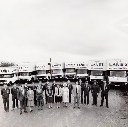 A black and white photo shows a group of men and women standing in a line outside in front of nine delivery trucks. The trucks are labeled "LANE'S OF CORNWALL." The people are dressed in various work and formal attire, smiling and facing the camera.