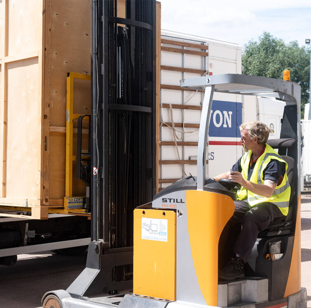 A worker wearing a high-visibility vest operates a forklift to unload a large wooden crate from a truck. The forklift is positioned in front of the truck, and the worker is focused on maneuvering the crate safely. Trees and additional trucks can be seen in the background.