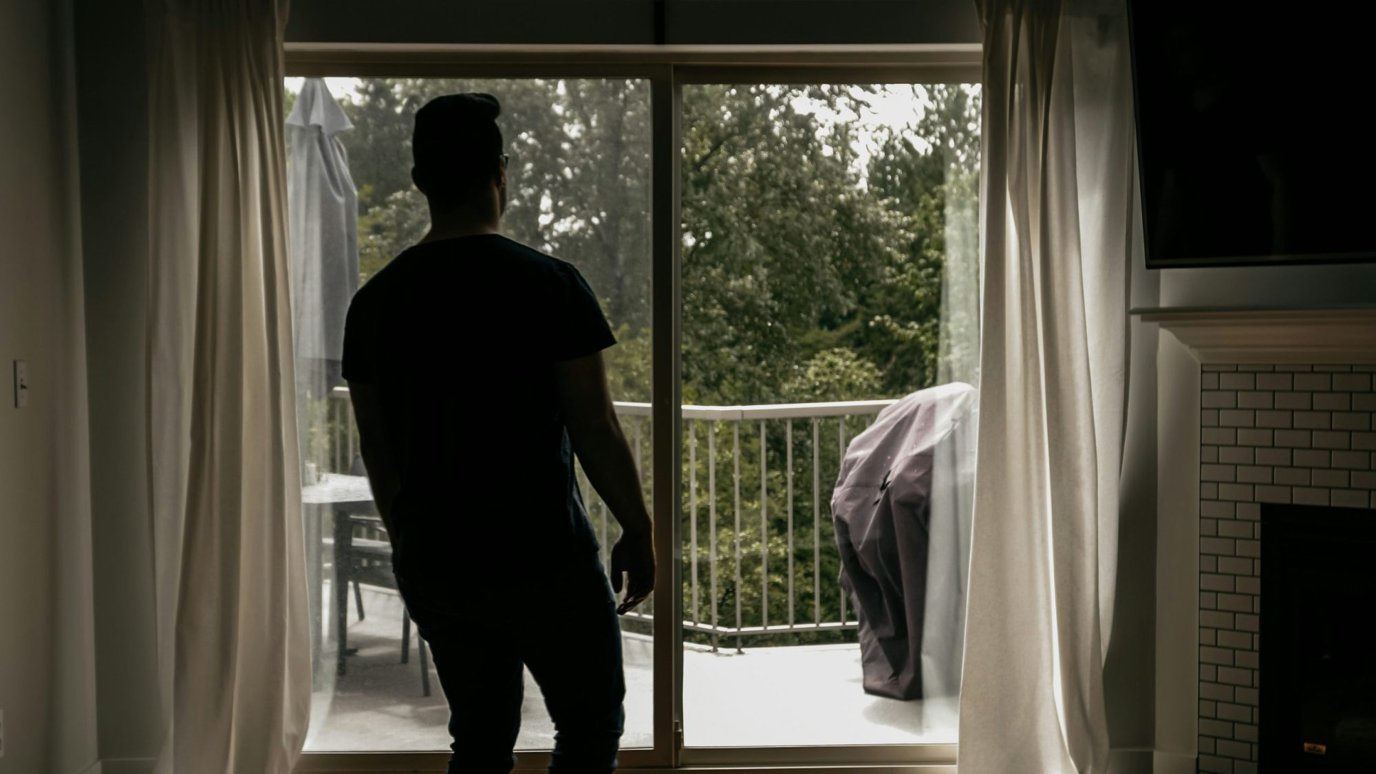 A silhouette of a person stands indoors, looking out through sliding glass doors towards a balcony with greenery outside. A covered object, possibly a grill, and patio furniture are visible on the balcony. Curtains frame the glass doors.